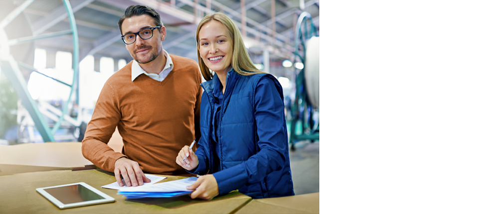 Personen arbeiten an einem Tisch mit Dokumenten und Tablet in einer Fabrikhalle, Überlagernd Text zur neuen ISO 9001-Norm.