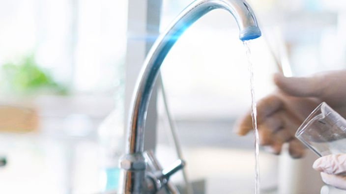 Wasser fließt aus einem Wasserhahn, während jemand mit Handschuhen ein Glas darunter hält.